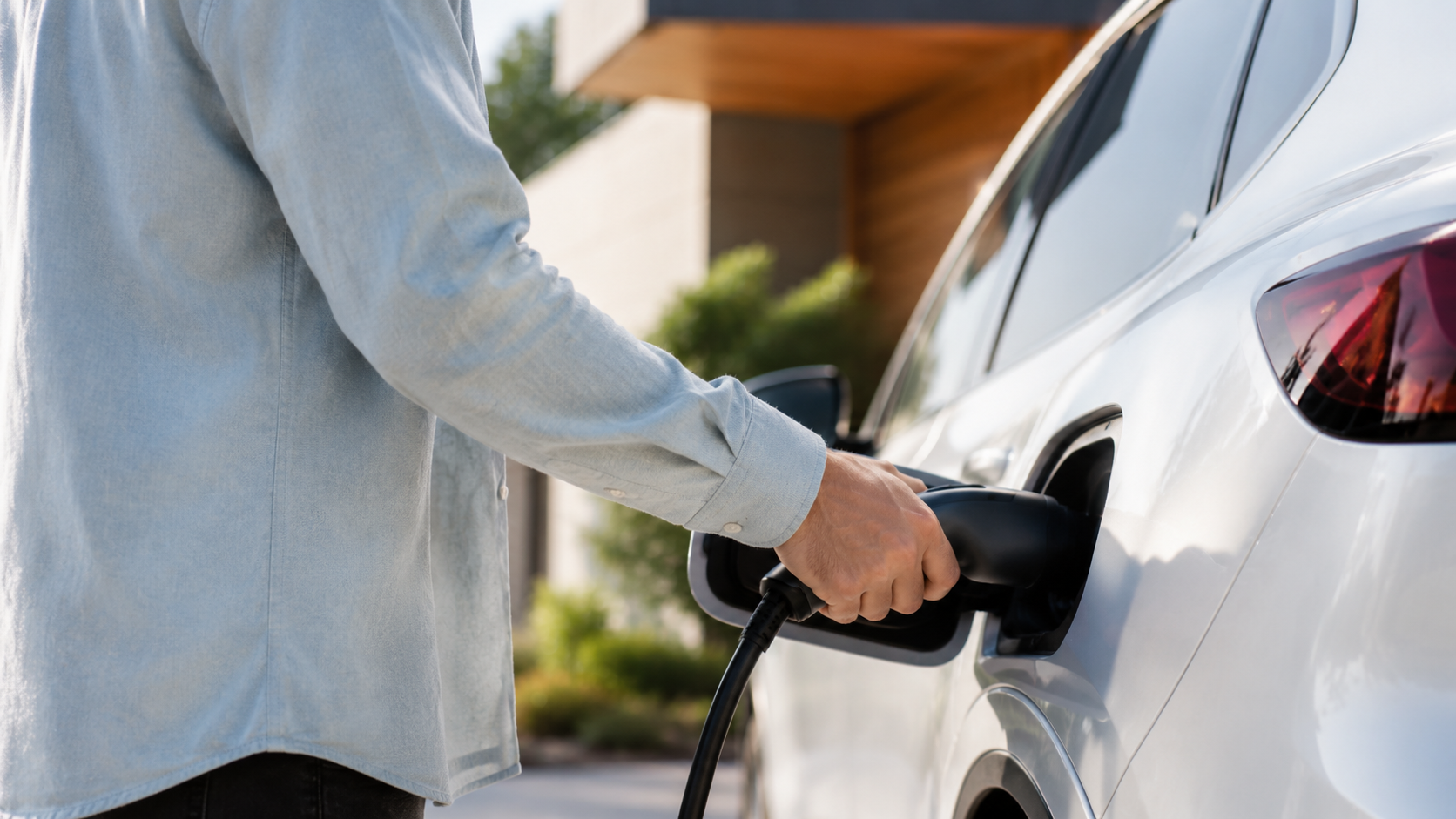Person in a light denim shirt plugs a charging cable into a white electric car in a driveway.